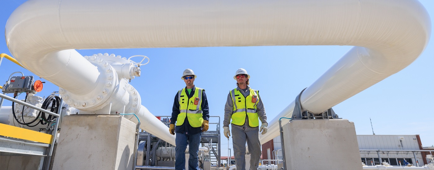 Two workers in safety gear walking near a white pipe