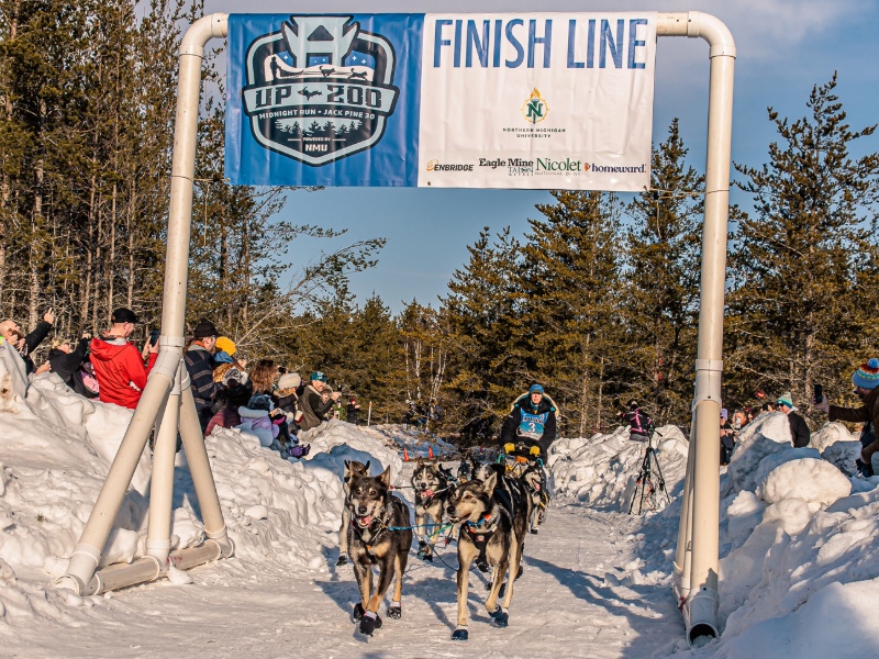 A musher crossing the finish line in dog sled with crowd on either side