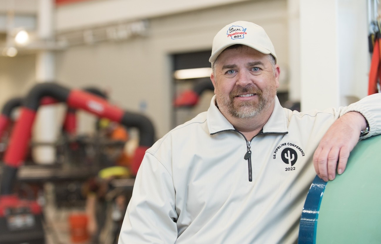Man in ball cap leaning on length of pipe in workshop