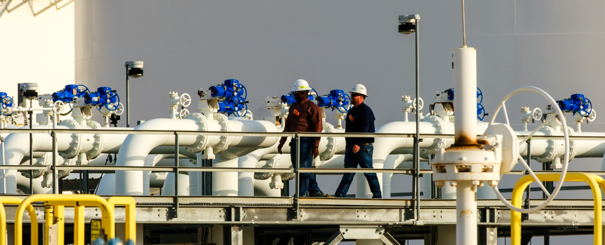 Workers walking on a manifold in front of a large tank